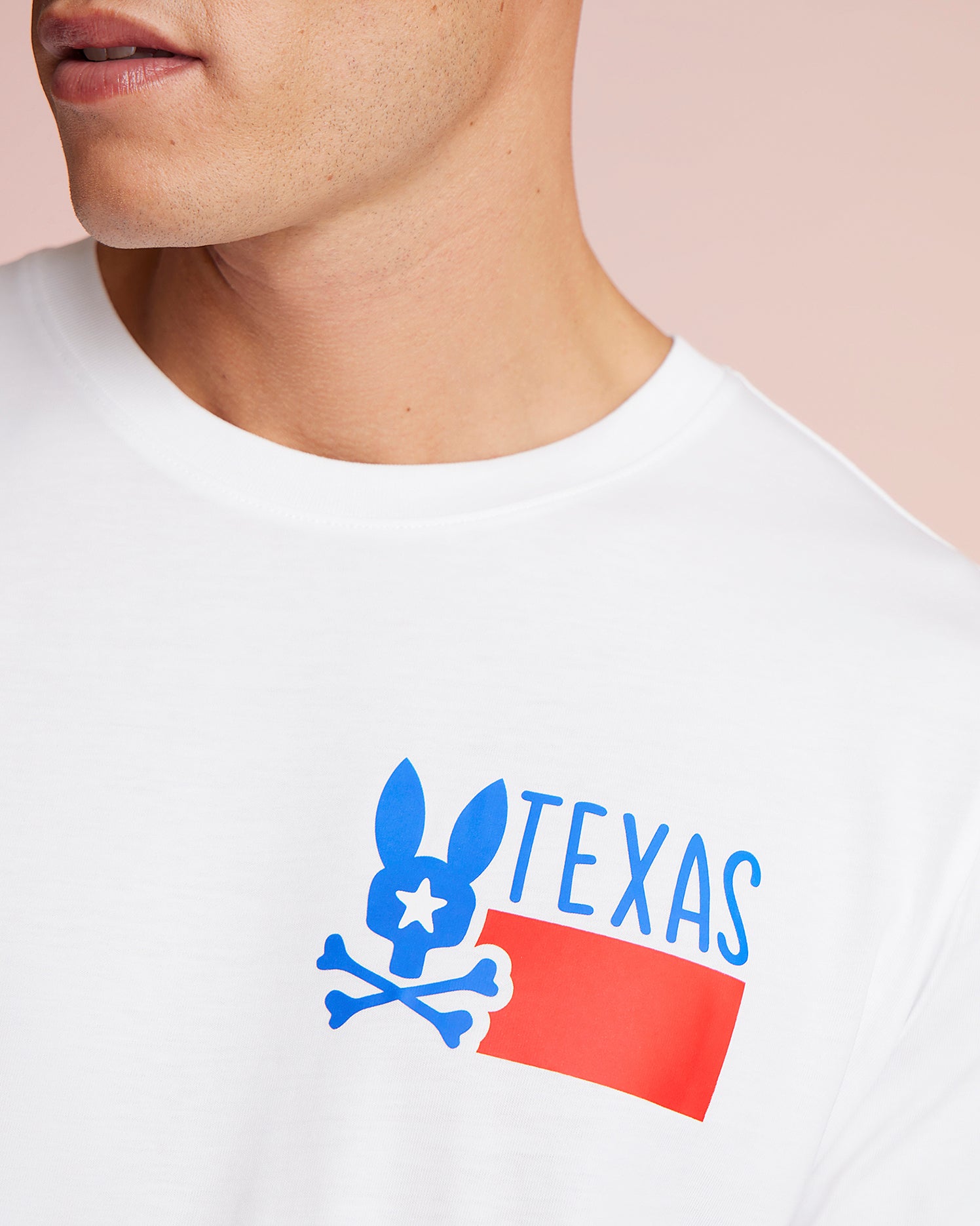 Close-up of a man wearing a white Psycho Bunny Pima cotton jersey T-shirt with a blue and red graphic of a stylized rabbit over the word "TEXAS." The background is a soft pink.