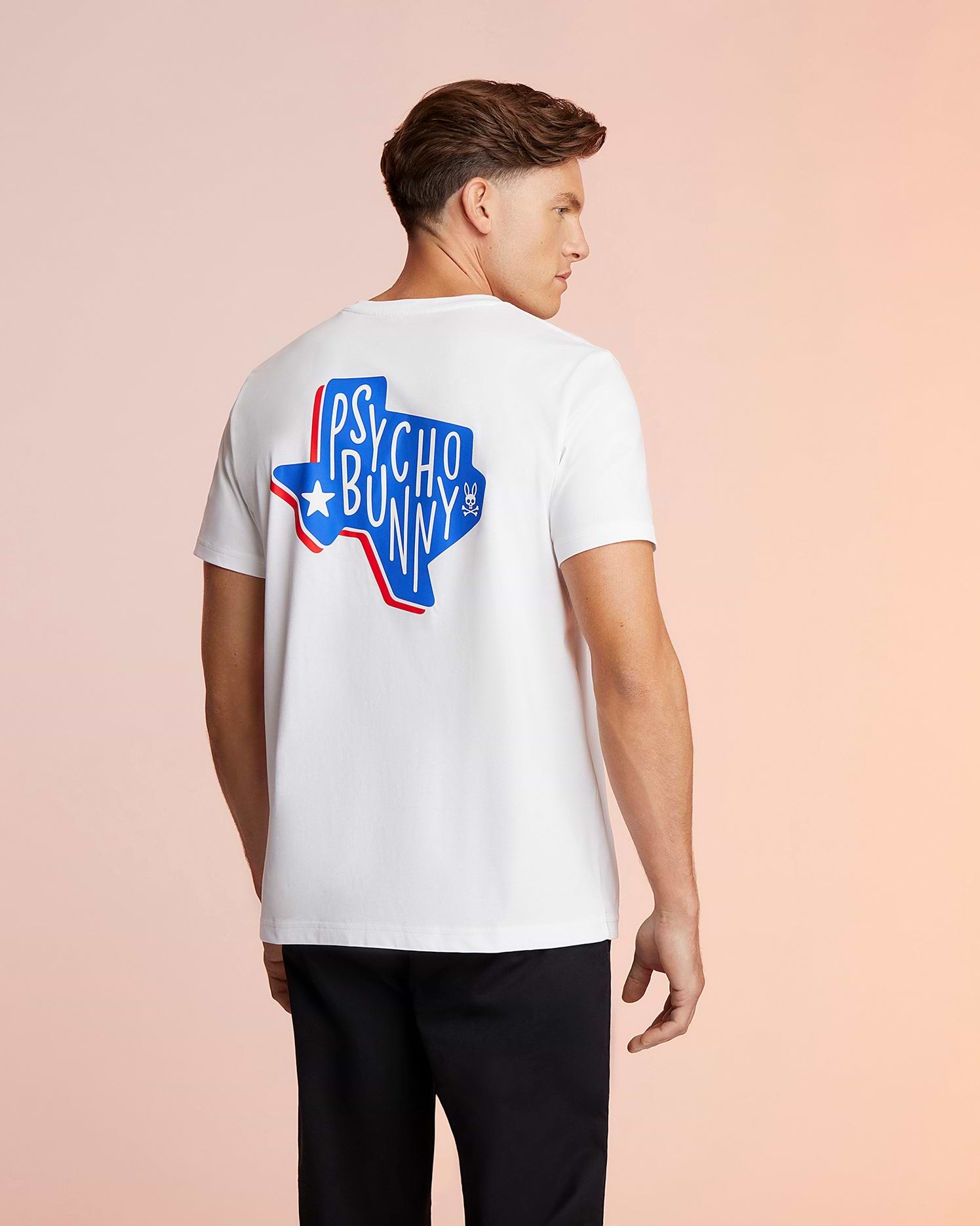 A man in a white Psycho Bunny Pima cotton jersey with a "Psycho Bunny" logo graphic in red and blue on the back, standing against a light peach background, looking to his left.