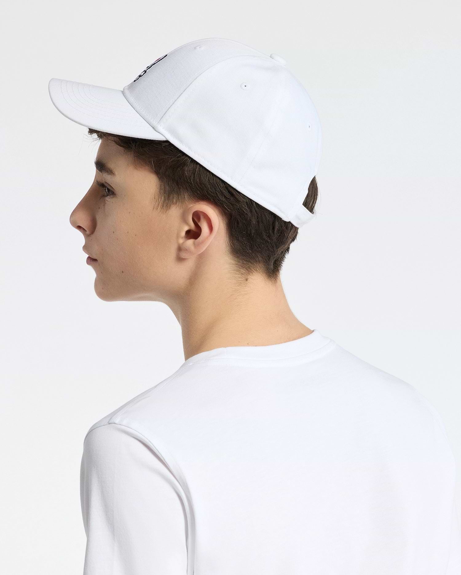 A person wearing a Psycho Bunny KIDS CLASSIC BASEBALL CAP in white, adorned with an embroidered bunny logo and featuring an adjustable fit, complements it with their white T-shirt. They are photographed from the side against a plain, light-colored backdrop.