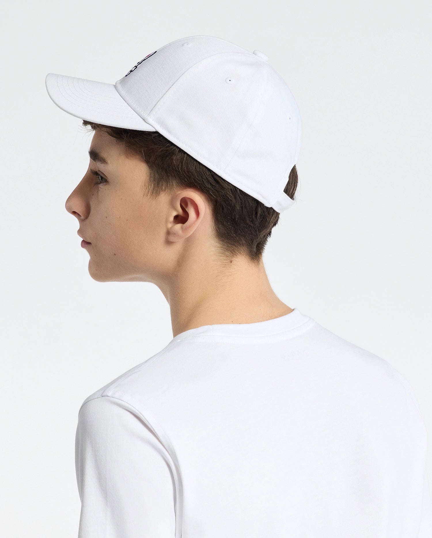 A person wearing a Psycho Bunny KIDS CLASSIC BASEBALL CAP in white, adorned with an embroidered bunny logo and featuring an adjustable fit, complements it with their white T-shirt. They are photographed from the side against a plain, light-colored backdrop.