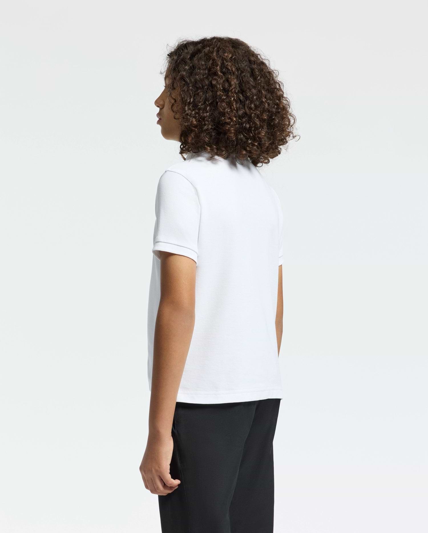 Curly-haired and facing away from the camera, a person wears a Psycho Bunny KIDS CLASSIC PIQUE POLO SHIRT in crisp white, paired with black pants, against a plain white background.