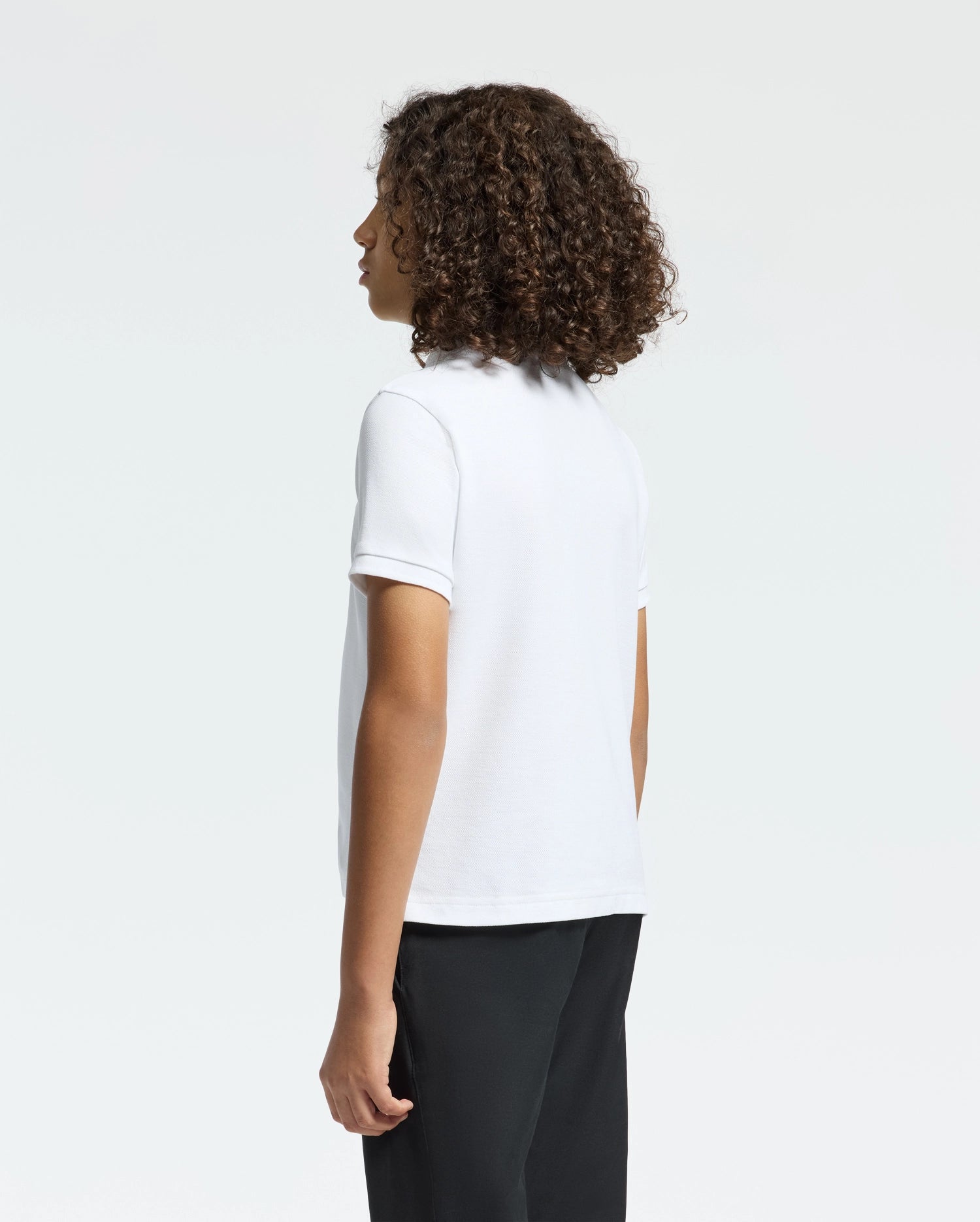 Curly-haired and facing away from the camera, a person wears a Psycho Bunny KIDS CLASSIC PIQUE POLO SHIRT in crisp white, paired with black pants, against a plain white background.