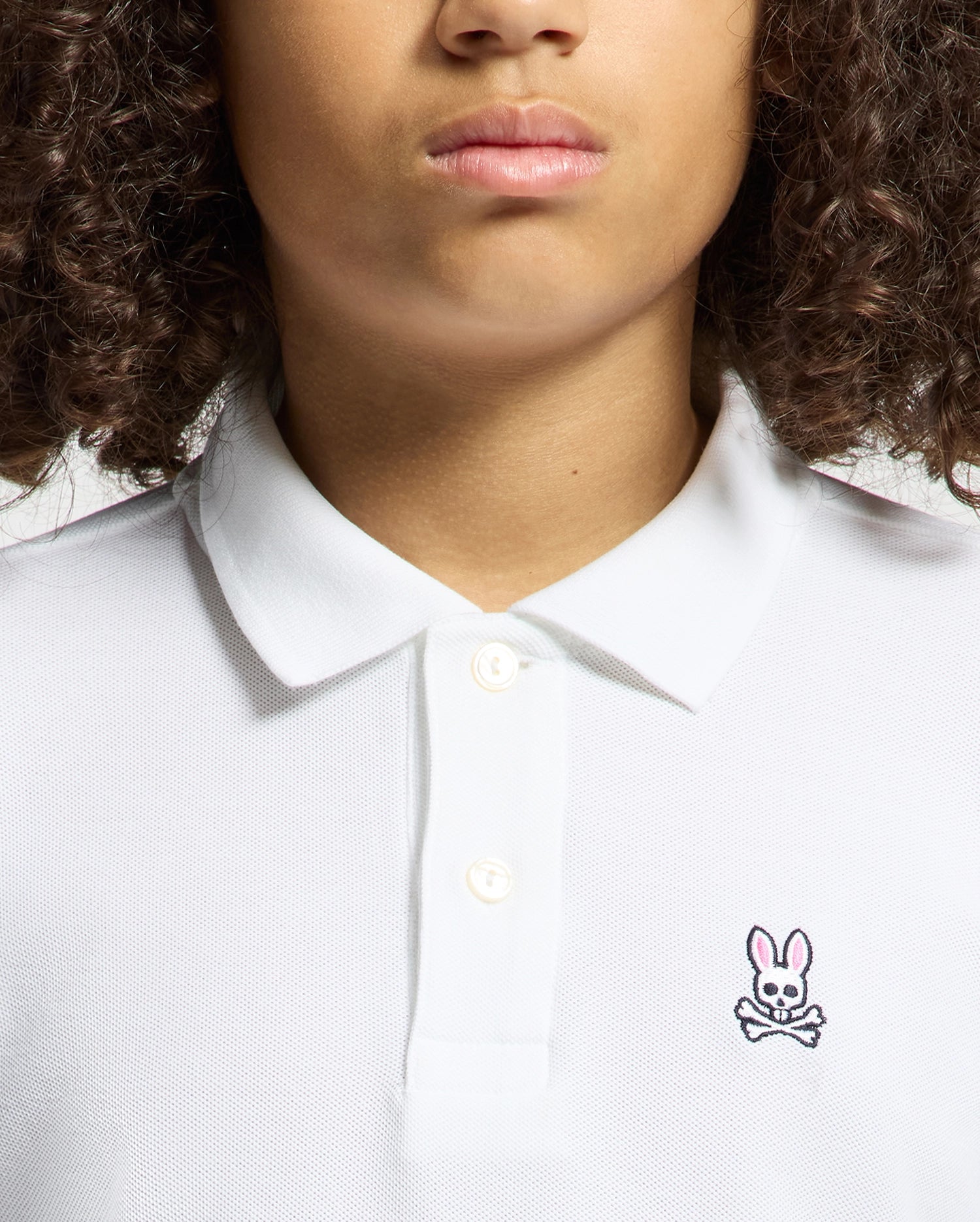 A curly-haired person wears a KIDS CLASSIC PIQUE POLO SHIRT by Psycho Bunny, ideal for back-to-school outfits. This white shirt stands out with its unique chest logo—a pink-eared rabbit above crossed bones.