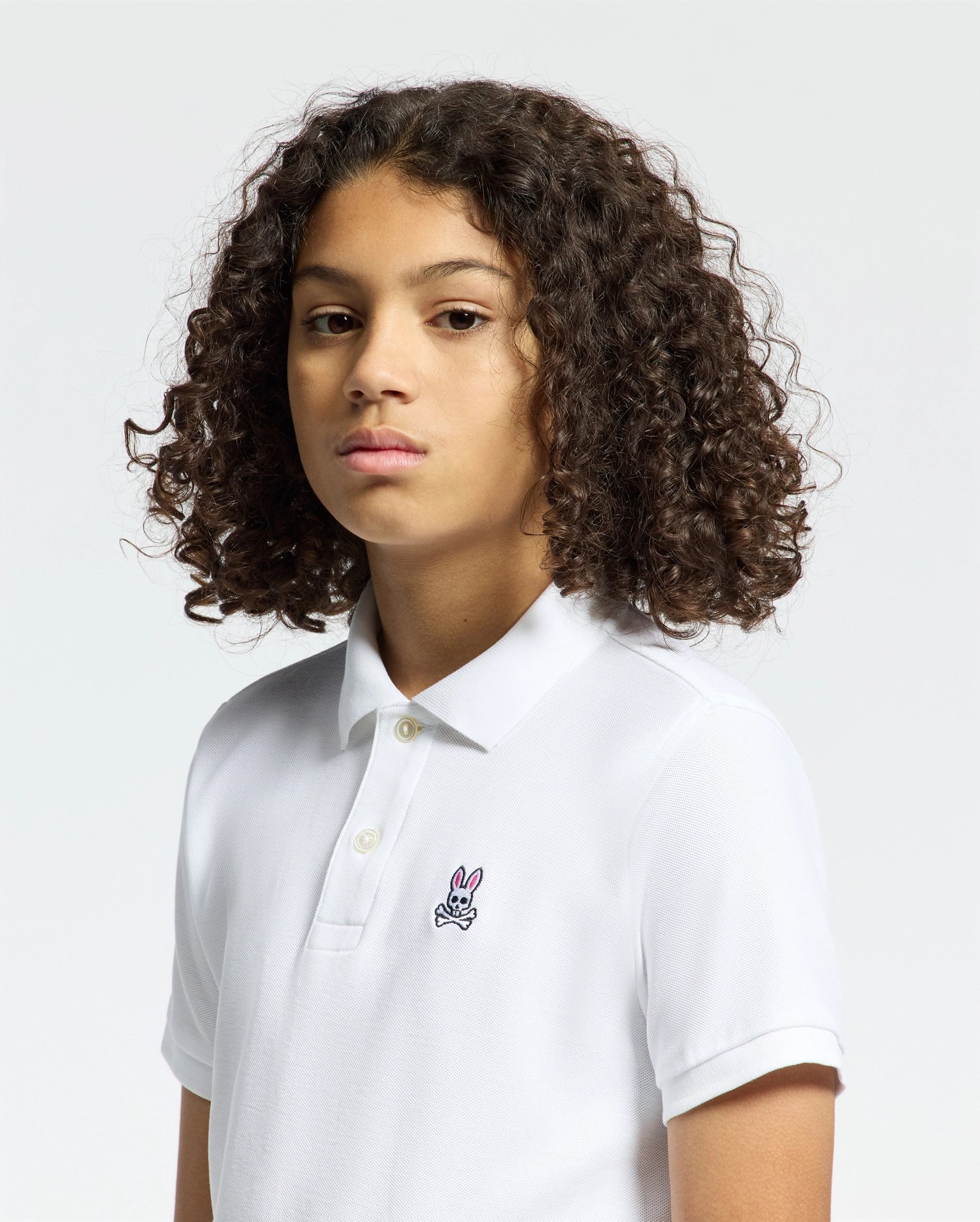 A person with curly hair wears a Psycho Bunny Kids Classic Pique Polo Shirt (B0K001CRPC), featuring a small embroidered logo on the chest. The plain, light background highlights this timeless choice, ideal for back-to-school outfits.
