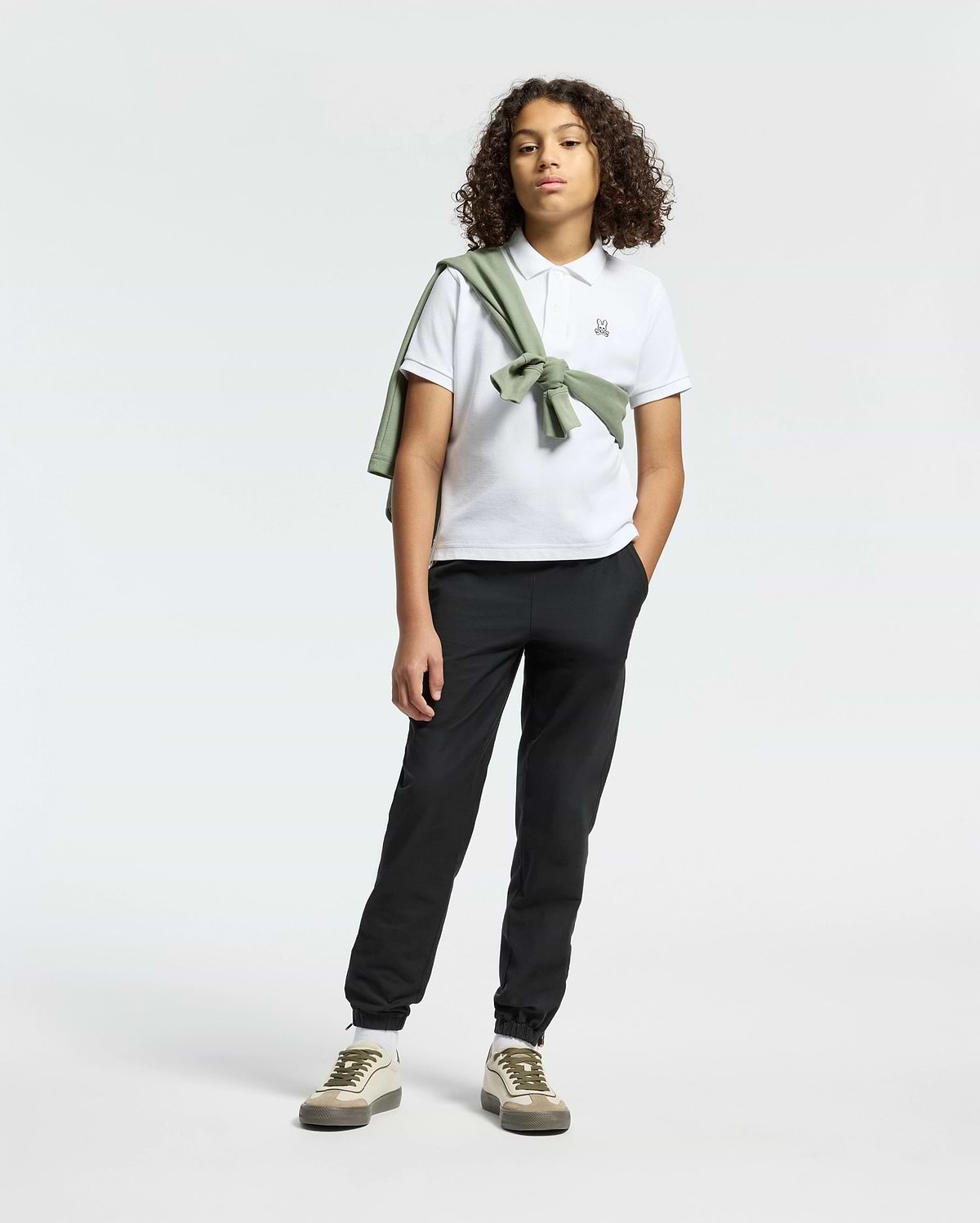 A teen confidently poses in Psycho Bunny’s KIDS CLASSIC PIQUE POLO SHIRT, paired with black pants. A green sweater drapes over their shoulder, adding back-to-school flair. With curly hair and sneakers, they stand against a neutral backdrop, ready for any occasion.
