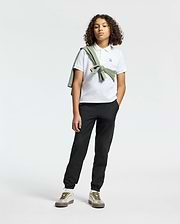 A teen confidently poses in Psycho Bunny’s KIDS CLASSIC PIQUE POLO SHIRT, paired with black pants. A green sweater drapes over their shoulder, adding back-to-school flair. With curly hair and sneakers, they stand against a neutral backdrop, ready for any occasion.