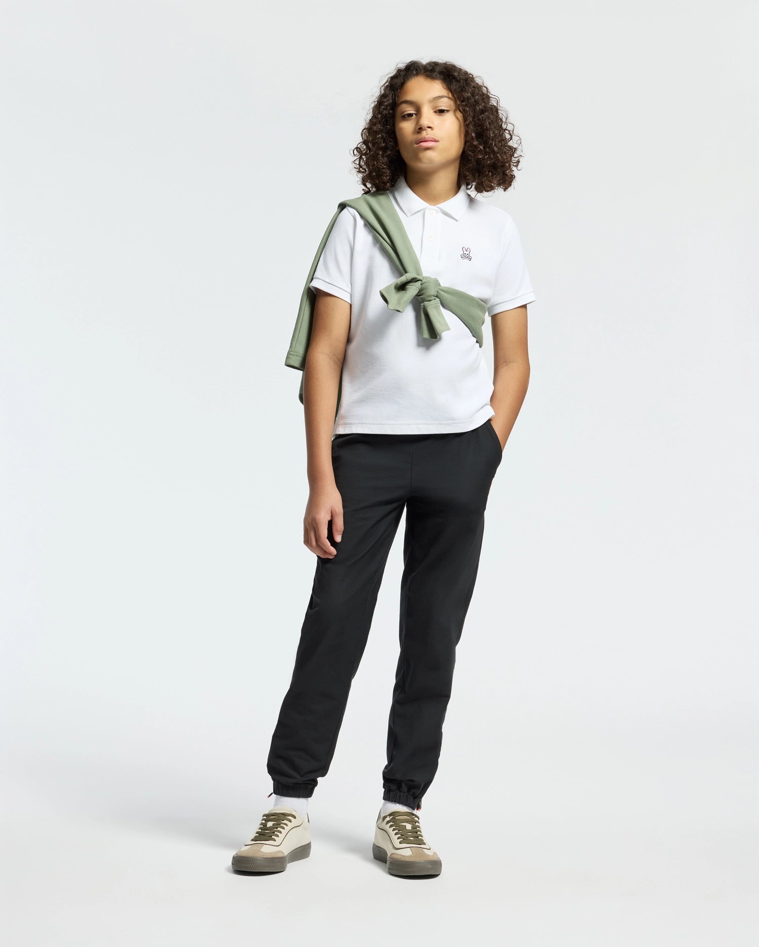 A teen confidently poses in Psycho Bunny’s KIDS CLASSIC PIQUE POLO SHIRT, paired with black pants. A green sweater drapes over their shoulder, adding back-to-school flair. With curly hair and sneakers, they stand against a neutral backdrop, ready for any occasion.