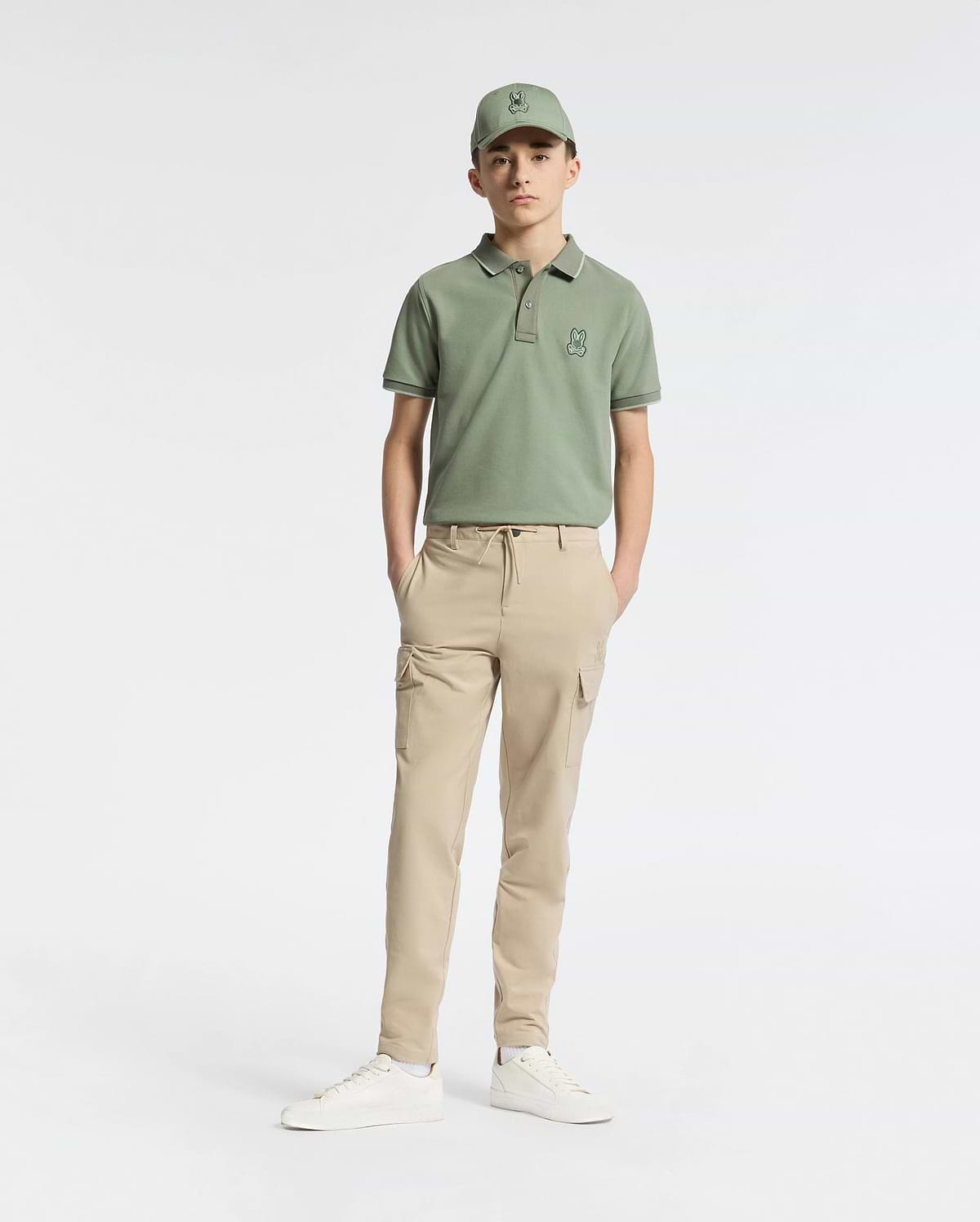 A teen wearing a green cap and matching polo shirt features the Psycho Bunny KIDS GREEN VALLEY CARGO PANT - B0P199E200, known for its cargo pockets and comfy light beige fabric, set against a white background. The outfit is completed with white sneakers and hands in pockets as he faces forward.