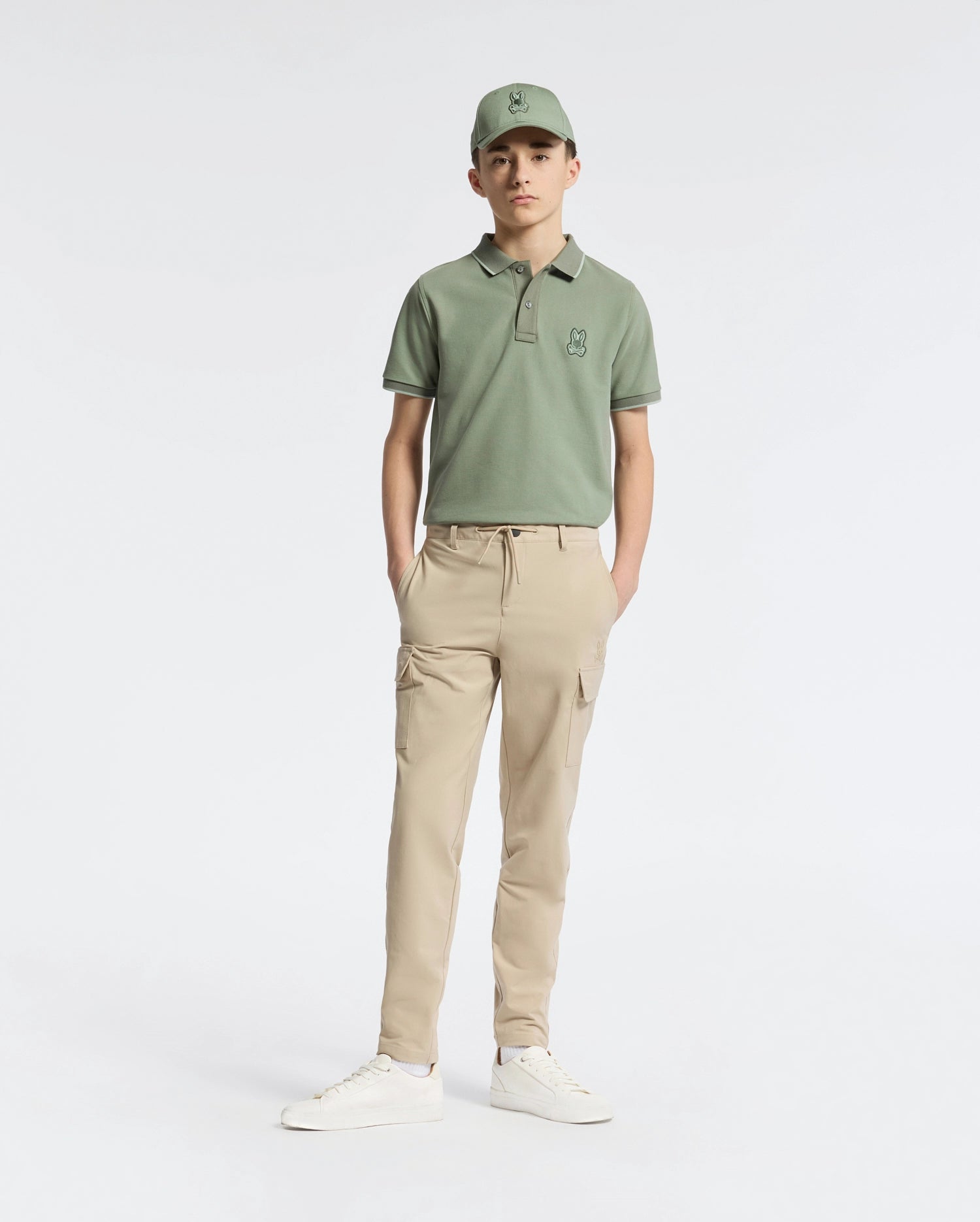 A teen wearing a green cap and matching polo shirt features the Psycho Bunny KIDS GREEN VALLEY CARGO PANT - B0P199E200, known for its cargo pockets and comfy light beige fabric, set against a white background. The outfit is completed with white sneakers and hands in pockets as he faces forward.