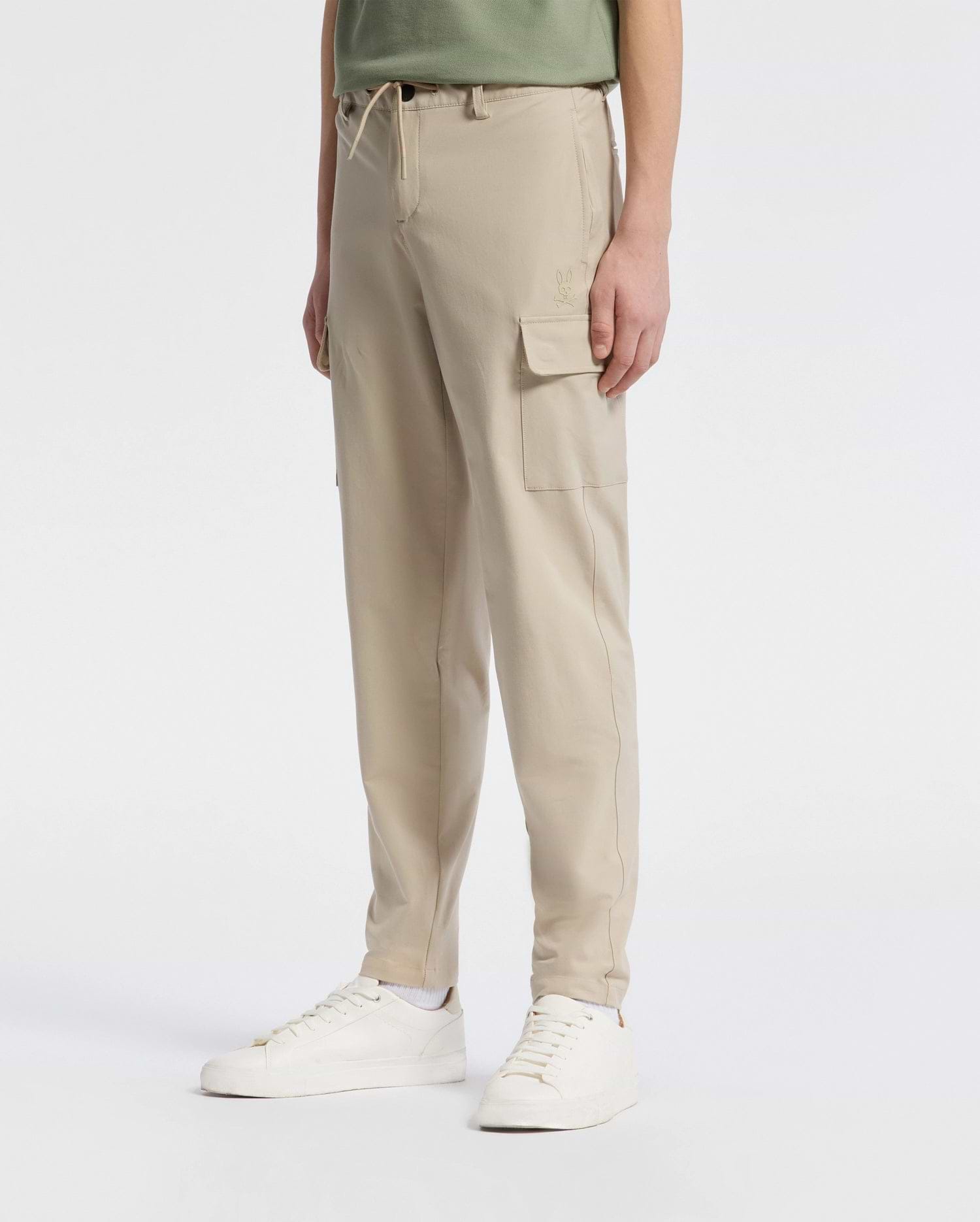 A person wears Psycho Bunny's KIDS GREEN VALLEY CARGO PANT - B0P199E200 and a green shirt against a white backdrop. The cargo pants boast pockets and a drawstring waist for comfort, paired with white sneakers.