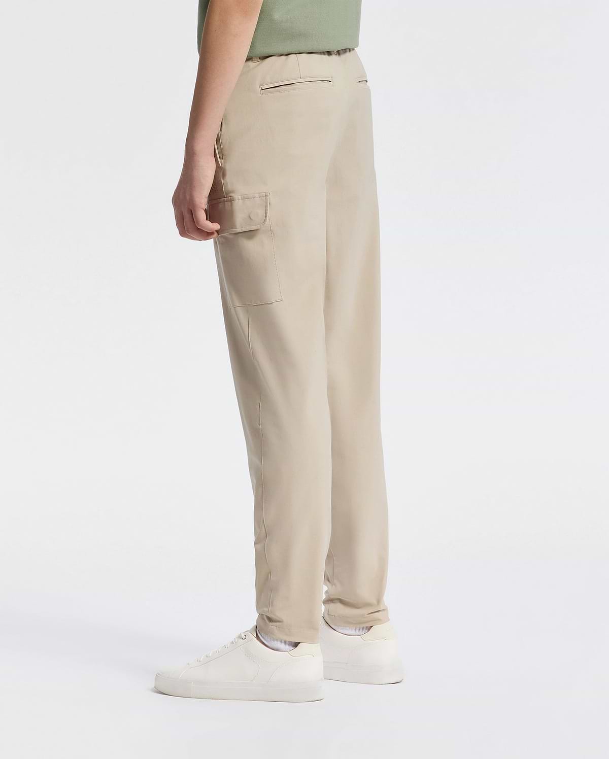 A person wearing Psycho Bunny's KIDS GREEN VALLEY CARGO PANT - B0P199E200 in beige and white sneakers stands against a plain background. These pants offer a stylish yet functional design with their comfortable fit and ample cargo pockets. They wear a light green top, partially visible.