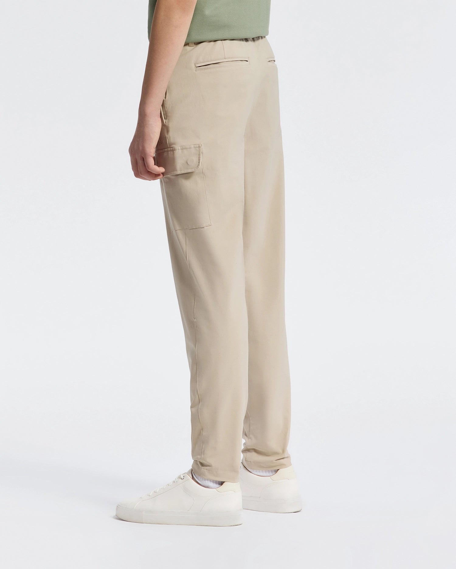 A person wearing Psycho Bunny's KIDS GREEN VALLEY CARGO PANT - B0P199E200 in beige and white sneakers stands against a plain background. These pants offer a stylish yet functional design with their comfortable fit and ample cargo pockets. They wear a light green top, partially visible.