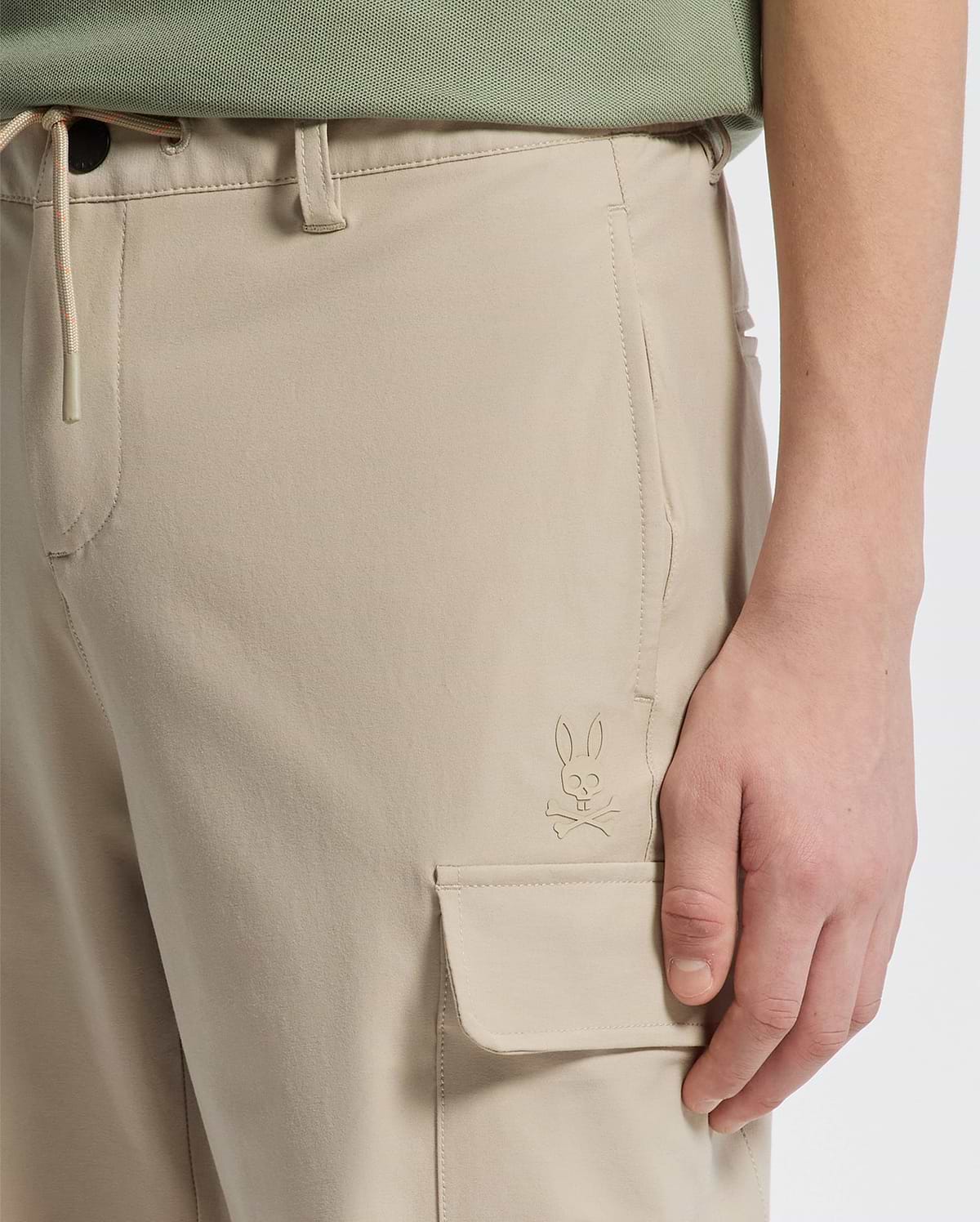 A close-up shows a person in Psycho Bunny's KIDS GREEN VALLEY CARGO PANT (B0P199E200) with a rabbit emblem on the pocket. Their hand rests nearby, paired with a green shirt for a comfortable fit.