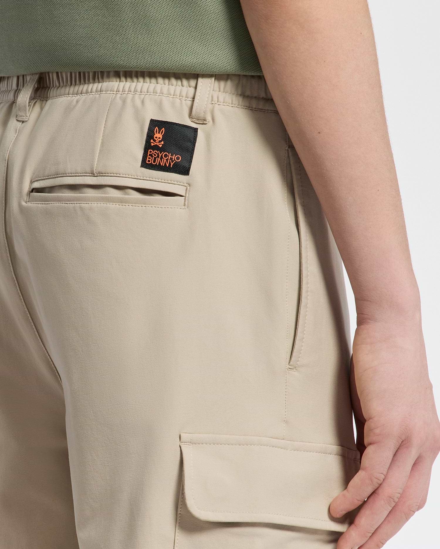 Someone wearing KIDS GREEN VALLEY CARGO PANT - B0P199E200 by Psycho Bunny, featuring an orange and black patch with a stylized rabbit and text on the back pocket. These comfortable-fit pants have side pockets, highlighting their rear design.