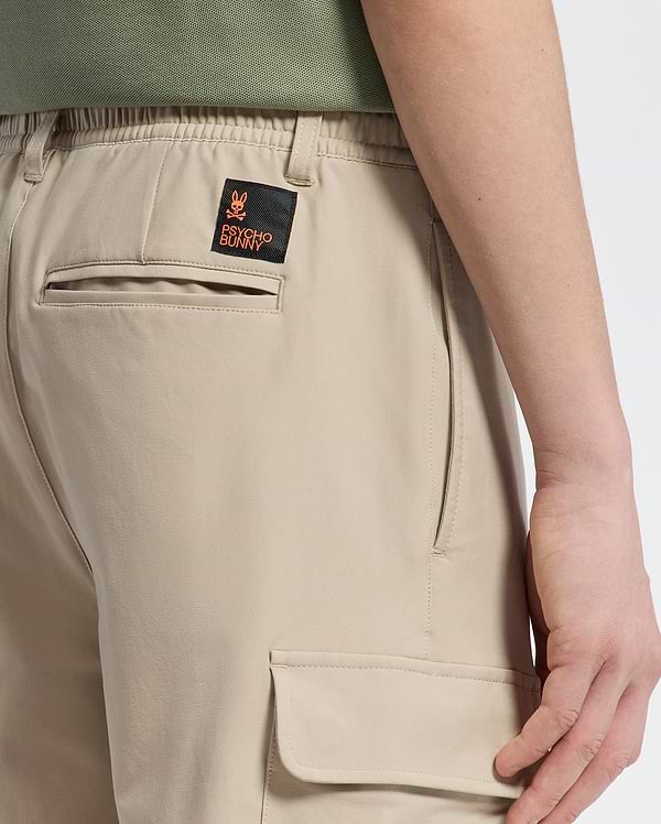 Someone wearing KIDS GREEN VALLEY CARGO PANT - B0P199E200 by Psycho Bunny, featuring an orange and black patch with a stylized rabbit and text on the back pocket. These comfortable-fit pants have side pockets, highlighting their rear design.