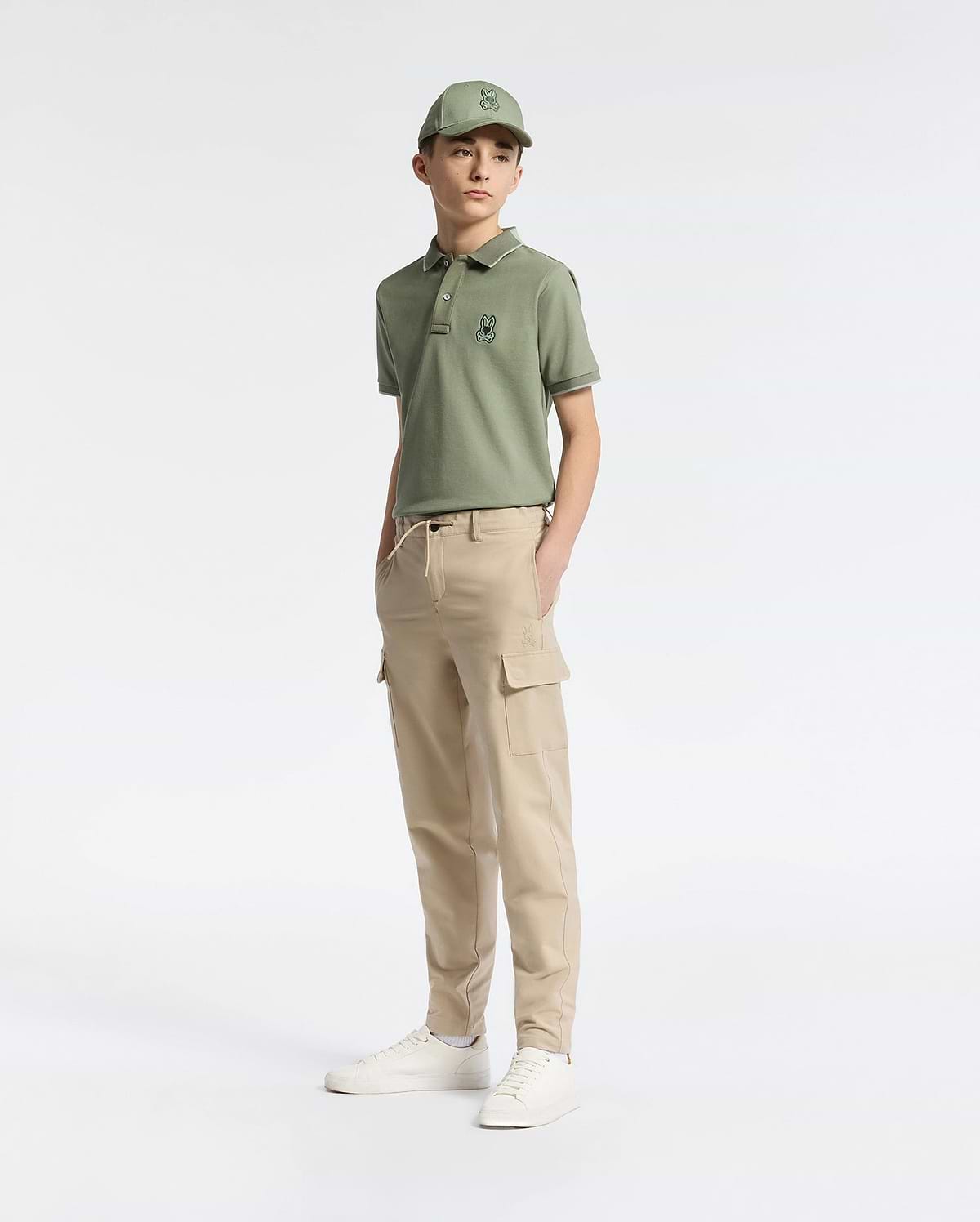 A teen boy in a green cap and polo with an embroidered logo wears Psycho Bunny's KIDS GREEN VALLEY CARGO PANT - B0P199E200 paired with white sneakers. The comfy fit and cargo pockets add to the casual, sporty vibe as he stands against a plain white background, gazing slightly to the side.