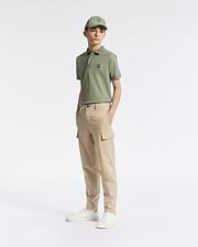 A teen boy in a green cap and polo with an embroidered logo wears Psycho Bunny's KIDS GREEN VALLEY CARGO PANT - B0P199E200 paired with white sneakers. The comfy fit and cargo pockets add to the casual, sporty vibe as he stands against a plain white background, gazing slightly to the side.