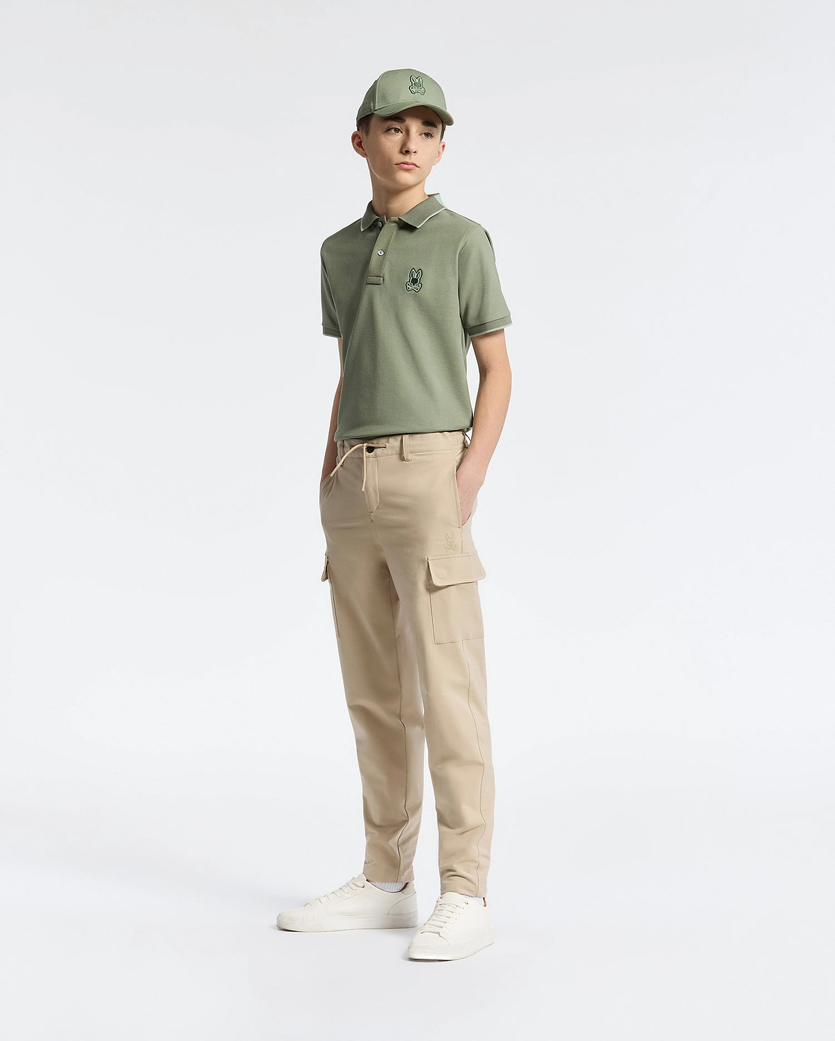A teen boy in a green cap and polo with an embroidered logo wears Psycho Bunny's KIDS GREEN VALLEY CARGO PANT - B0P199E200 paired with white sneakers. The comfy fit and cargo pockets add to the casual, sporty vibe as he stands against a plain white background, gazing slightly to the side.