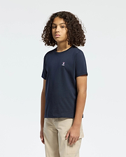 A curly-haired person wears a Psycho Bunny Kids Classic Crew Neck Tee in navy blue paired with beige pants, set against a plain white background. The 100% Pima cotton tee sports an emblem on the chest, ideal for back-to-school style.