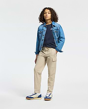 A person with curly hair wears a blue denim jacket over the KIDS CLASSIC CREW NECK TEE - B0U014CRPC by Psycho Bunny and tan cargo pants, creating a confident back-to-school look. They stand with one hand in their pocket and sport blue and white sneakers against a plain white background.