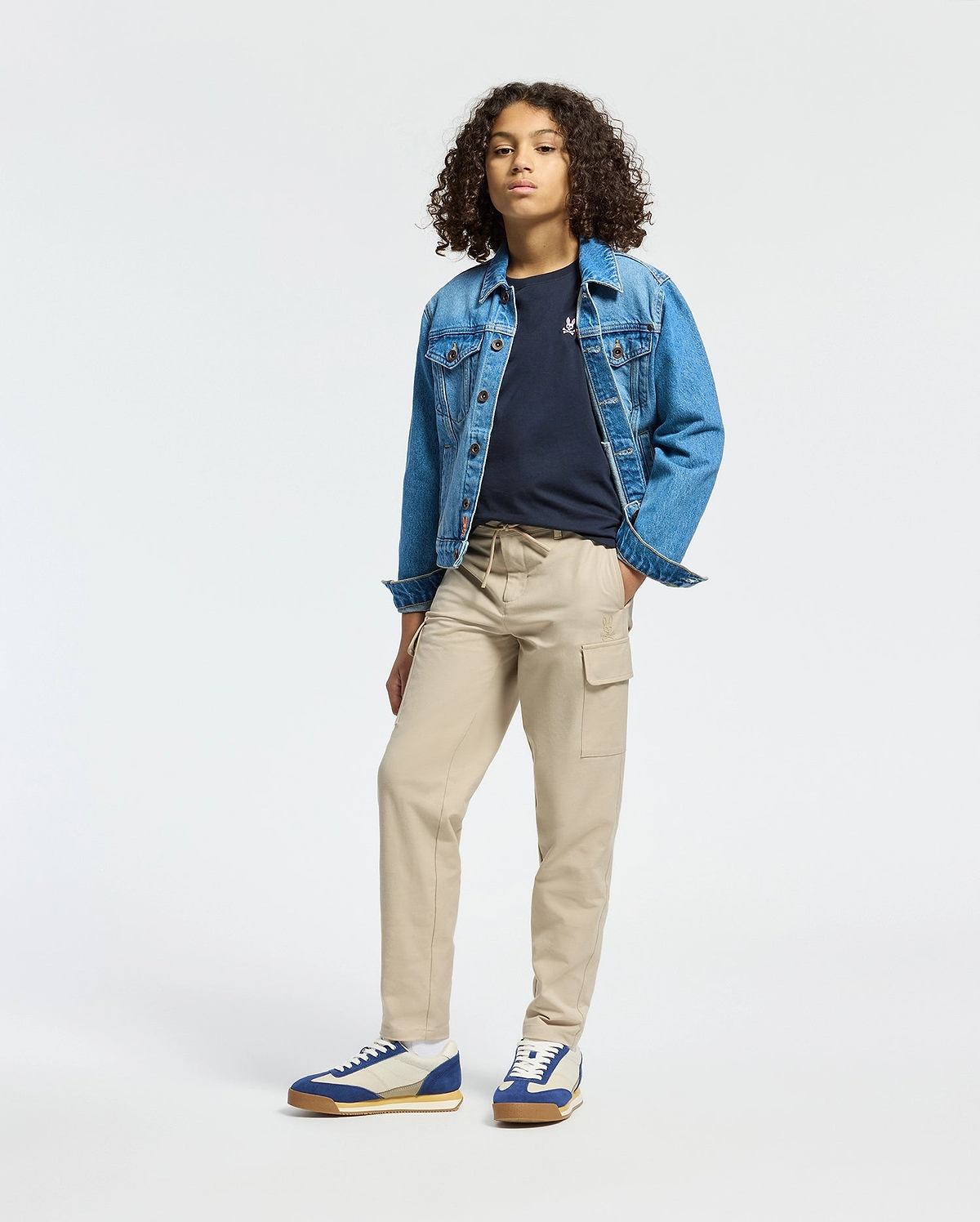 A person with curly hair wears a blue denim jacket over the KIDS CLASSIC CREW NECK TEE - B0U014CRPC by Psycho Bunny and tan cargo pants, creating a confident back-to-school look. They stand with one hand in their pocket and sport blue and white sneakers against a plain white background.