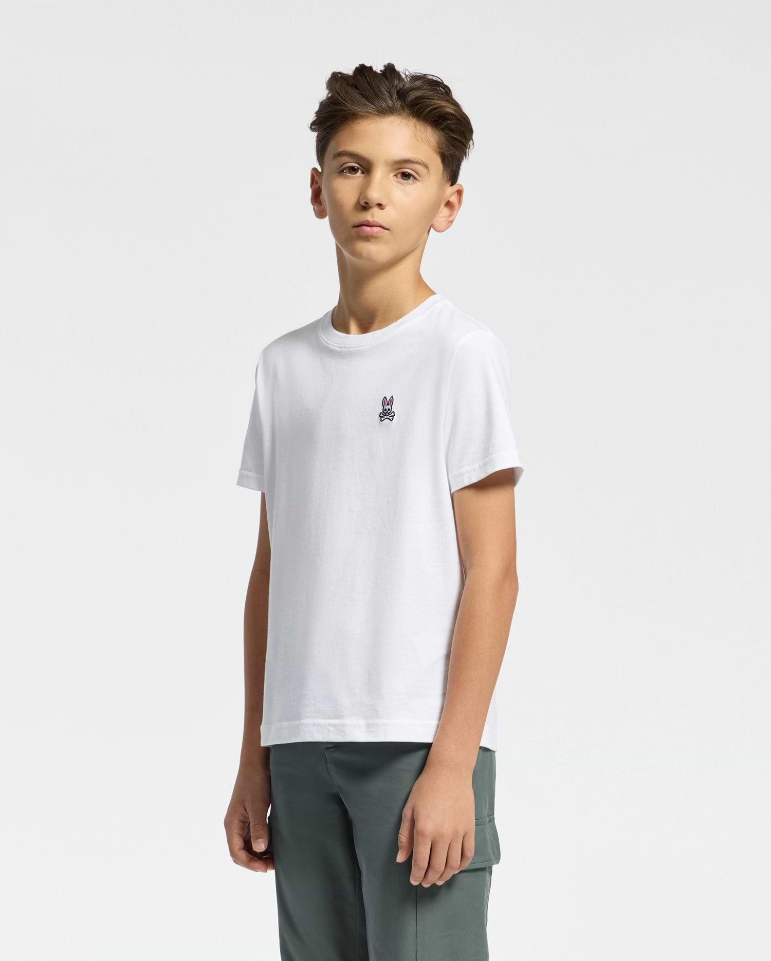 A young person with short brown hair wears Psycho Bunny's KIDS CLASSIC CREW NECK TEE - B0U014CRPC, crafted from 100% Pima cotton and featuring a chest graphic, paired with gray pants. Ideal for back-to-school outfits, they stand confidently against a plain white background.