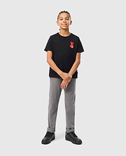 A young boy stands confidently against a light gray background, wearing the KIDS HAWAII CITY TEE from Psycho Bunny, made of soft Pima cotton. His black tee features a red bunny logo on the chest and is paired with gray pants and black sneakers. With braided hair and a slight smile, he clasps his hands loosely.