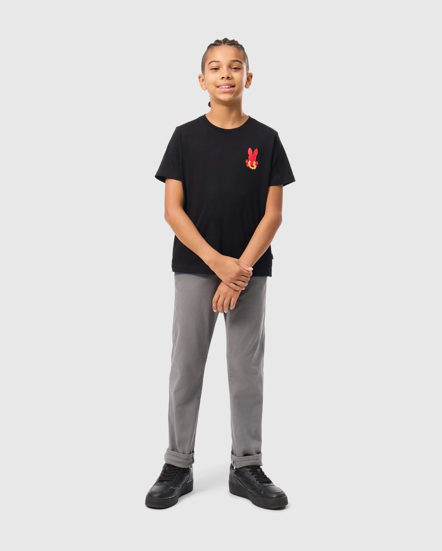 A young boy stands confidently against a light gray background, wearing the KIDS HAWAII CITY TEE from Psycho Bunny, made of soft Pima cotton. His black tee features a red bunny logo on the chest and is paired with gray pants and black sneakers. With braided hair and a slight smile, he clasps his hands loosely.
