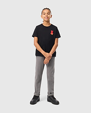 A young boy stands confidently against a light gray background, wearing the KIDS HAWAII CITY TEE from Psycho Bunny, made of soft Pima cotton. His black tee features a red bunny logo on the chest and is paired with gray pants and black sneakers. With braided hair and a slight smile, he clasps his hands loosely.