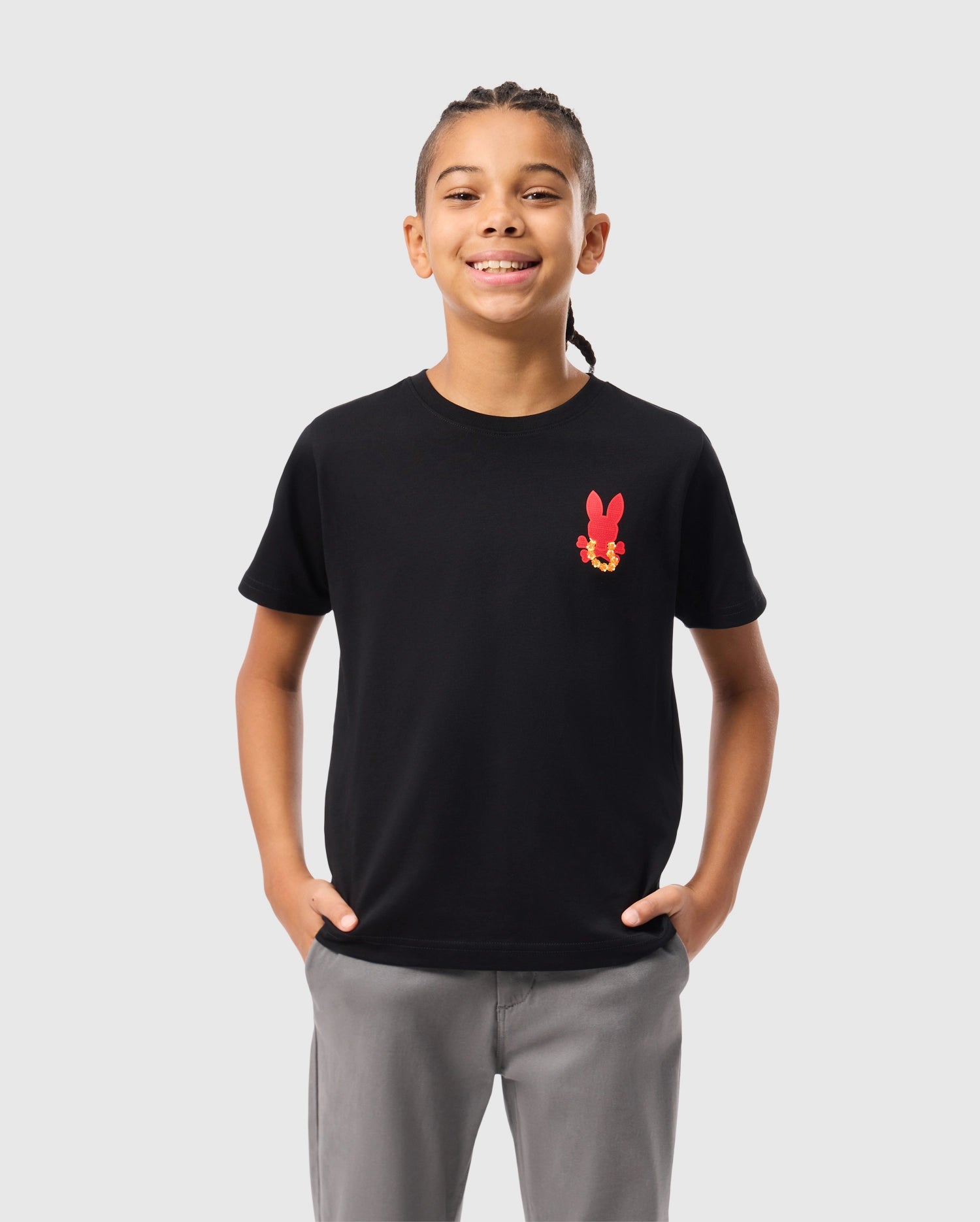 A child with braided hair is wearing the KIDS HAWAII CITY TEE from Psycho Bunny, a black Pima cotton t-shirt with a red bunny graphic, paired with gray pants. They stand hands in pockets against a plain white background.