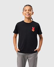 A child with braided hair is wearing the KIDS HAWAII CITY TEE from Psycho Bunny, a black Pima cotton t-shirt with a red bunny graphic, paired with gray pants. They stand hands in pockets against a plain white background.
