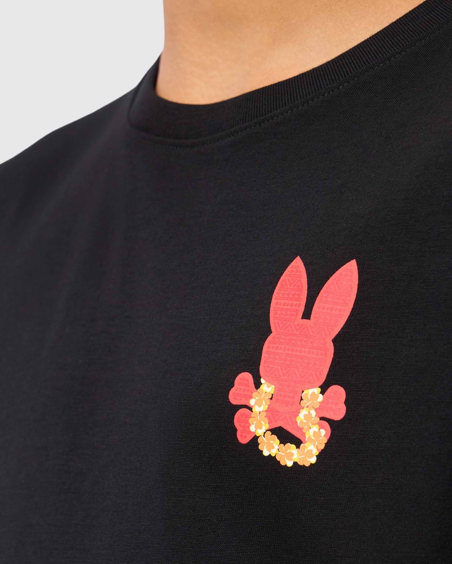 A person sports the Psycho Bunny KIDS HAWAII CITY TEE - B0U169Y1PC, crafted from Pima cotton in black, showcasing a red bunny graphic embellished with a chain of small yellow flowers, adding charm against the plain, light background.