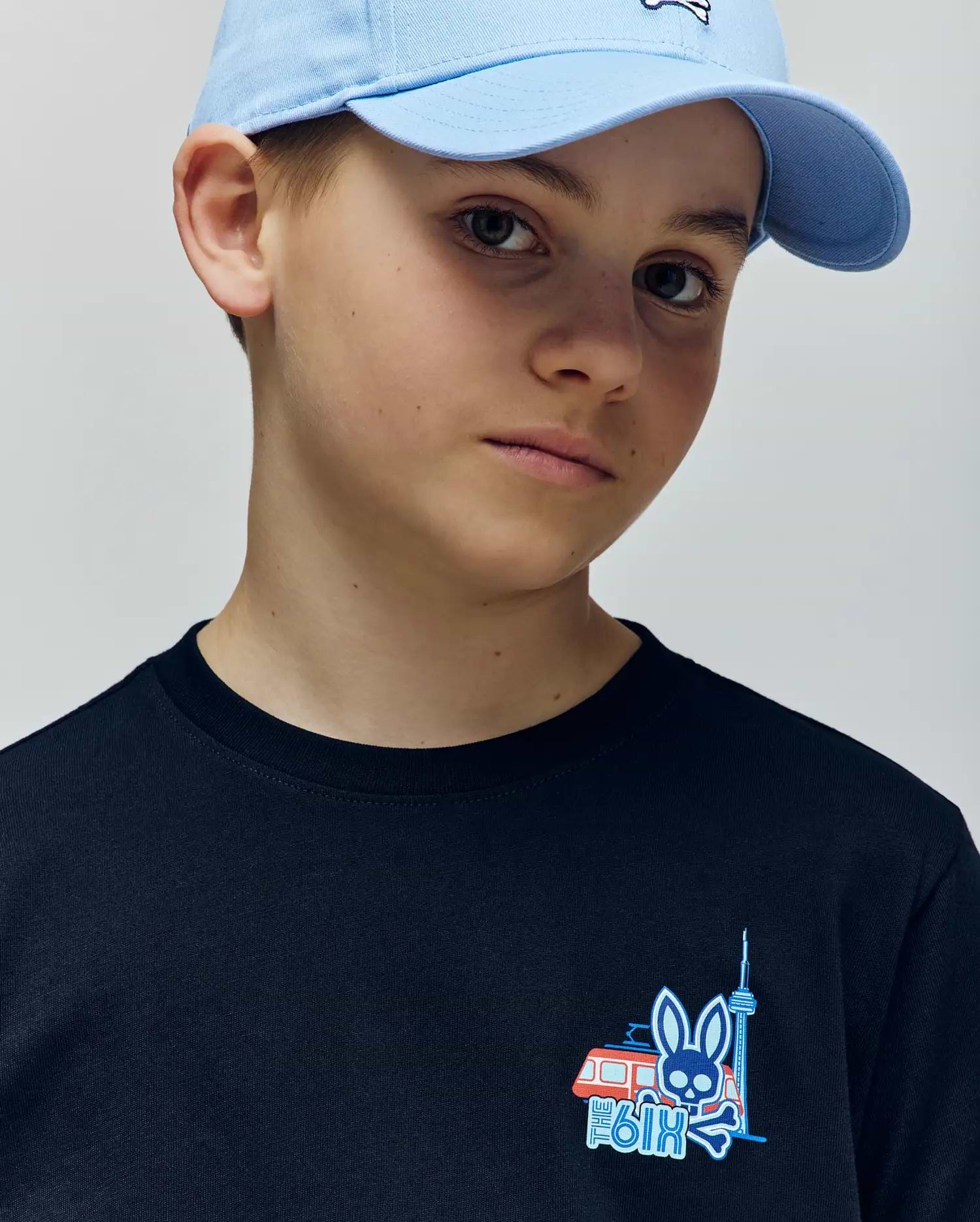 A young boy in a light blue cap and Psycho Bunny KIDS TORONTO TEE (B0U383F200), made of soft Pima cotton, featuring bold graphic prints—a blue bunny, red and white van, and tower—looks slightly to the side.