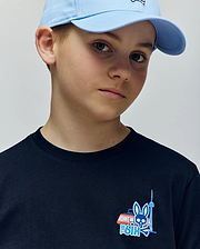 A young boy in a light blue cap and Psycho Bunny KIDS TORONTO TEE (B0U383F200), made of soft Pima cotton, featuring bold graphic prints—a blue bunny, red and white van, and tower—looks slightly to the side.