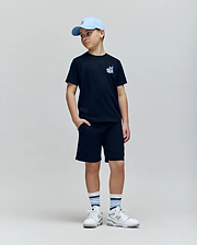 A boy in a light blue cap, black Psycho Bunny KIDS TORONTO TEE (B0U383F200), black shorts, white sneakers, and white striped socks stands with one hand in his pocket against a plain light background.