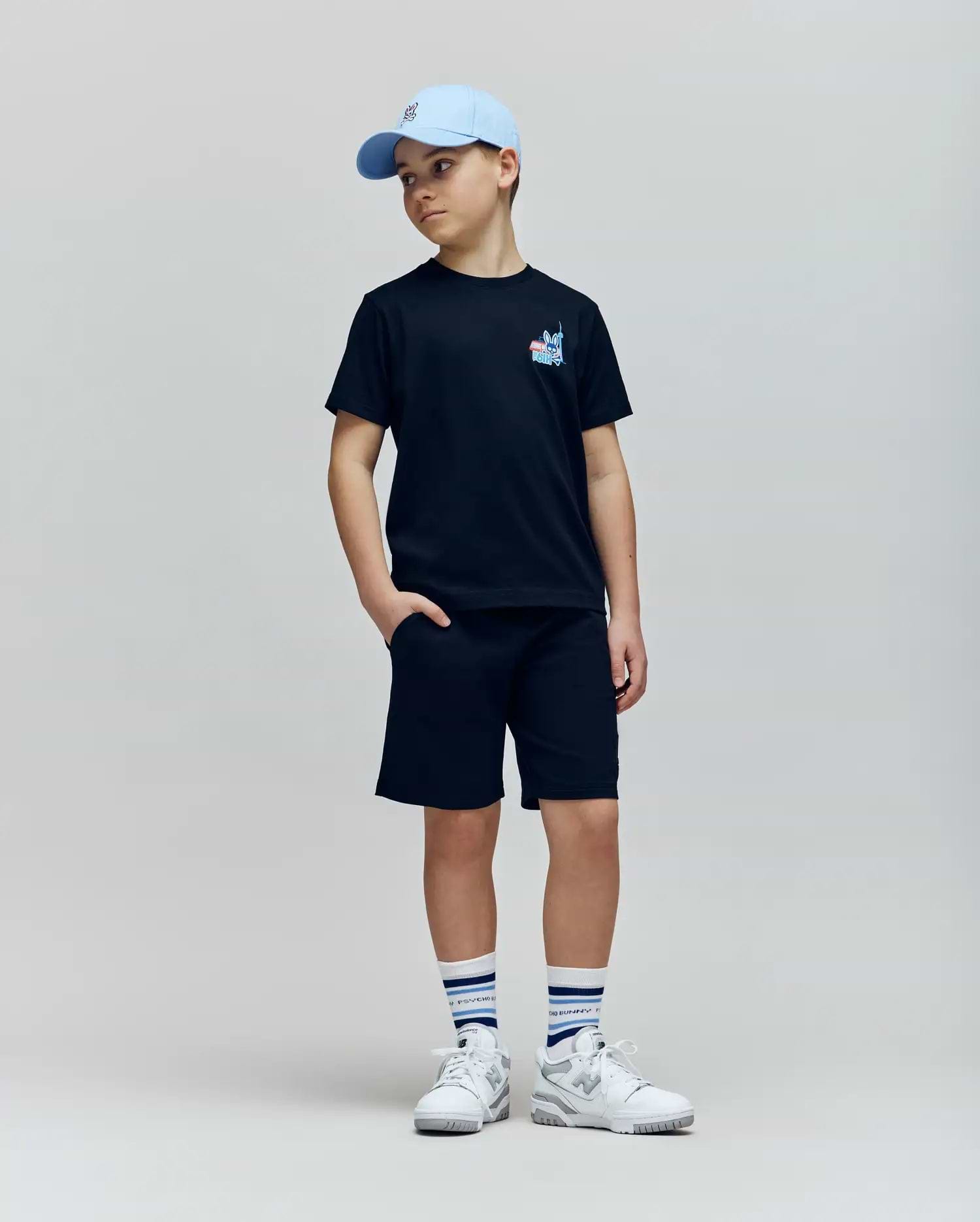 A boy in a light blue cap, black Psycho Bunny KIDS TORONTO TEE (B0U383F200), black shorts, white sneakers, and white striped socks stands with one hand in his pocket against a plain light background.