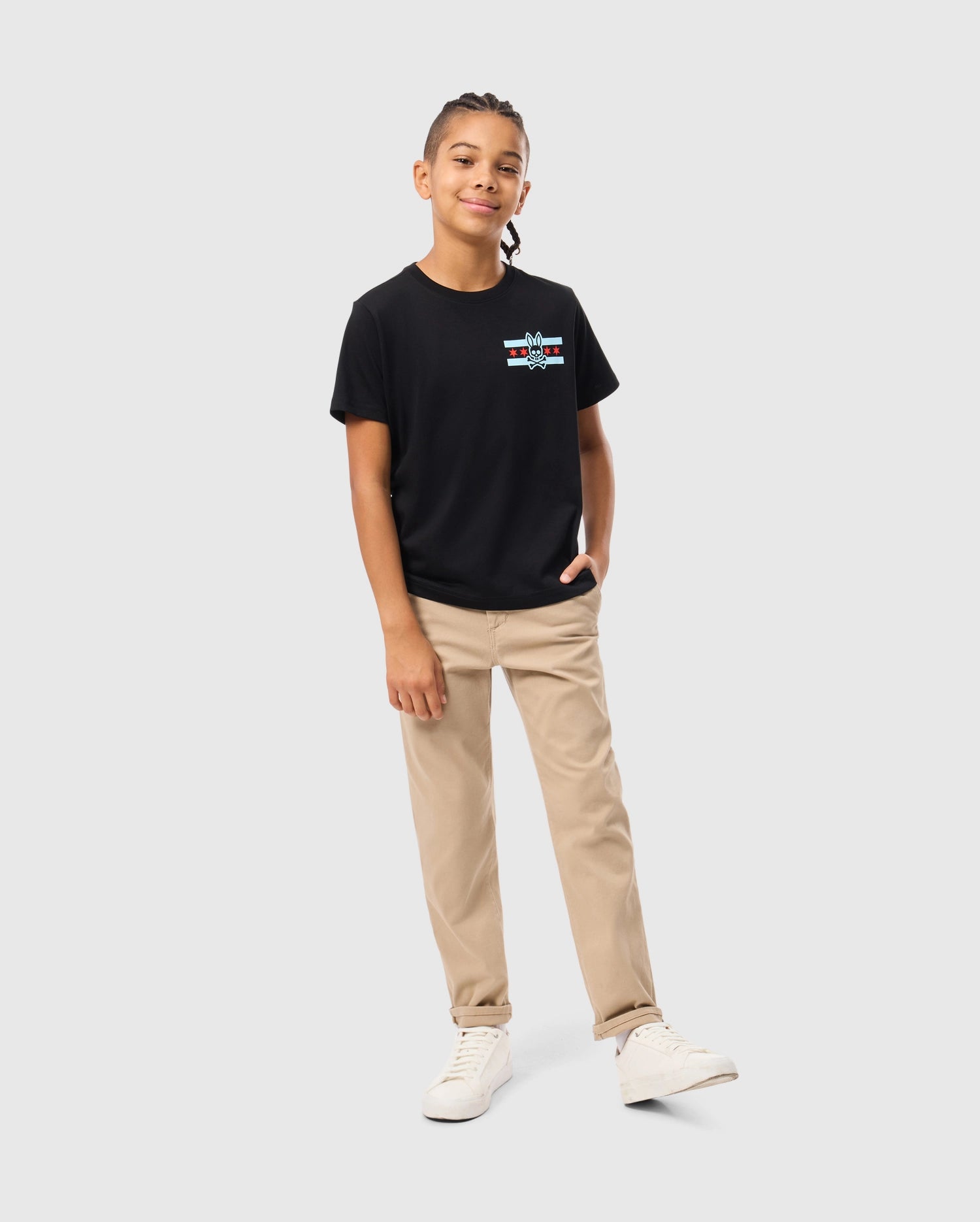A young person with long braided hair confidently stands against a light gray background, wearing the Psycho Bunny KIDS CHICAGO CITY TEE - B0U551W1PC in black, crafted from soft Pima cotton jersey, paired with beige pants and white sneakers, with one hand in their pocket.