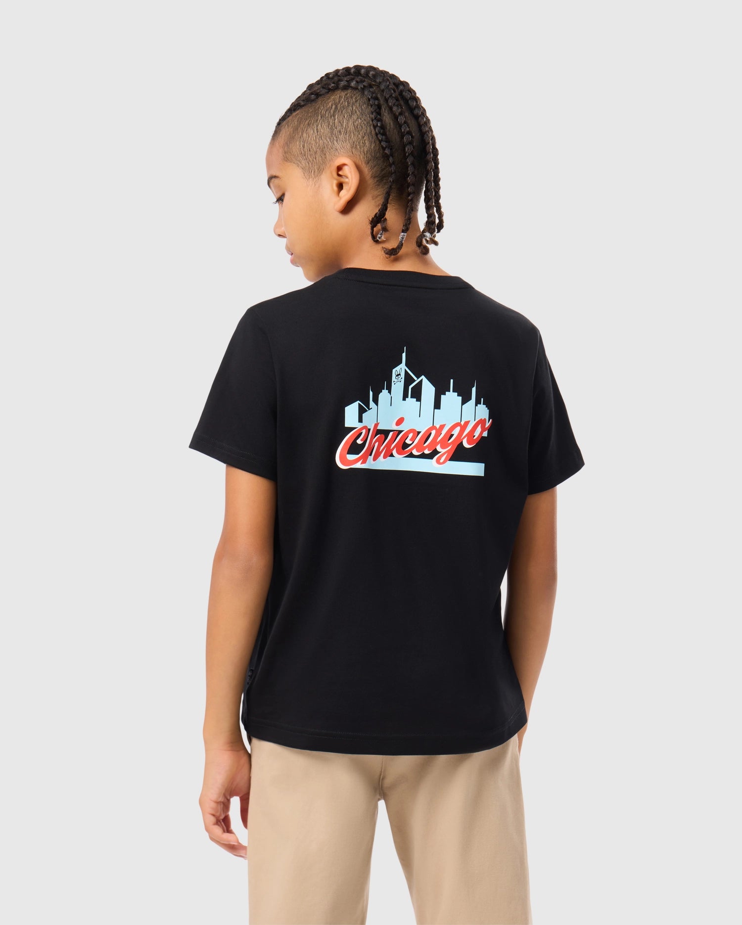 A child with braided hair stands facing away, wearing the Psycho Bunny KIDS CHICAGO CITY TEE - B0U551W1PC, a black t-shirt crafted from soft Pima cotton jersey and showcasing the word "Chicago" along with a skyline graphic. Light-colored pants complete this casual look.