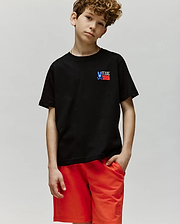 A boy with curly hair wears the Psycho Bunny KIDS TEXAS TEE (B0U553W1PC) in black, featuring a small "TEXAS" graphic, and red shorts. He stands against a light gray background, looking at the camera with a relaxed expression.