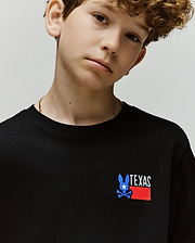 A young boy wears the Psycho Bunny KIDS TEXAS TEE (B0U553W1PC), a black Pima cotton shirt with a blue bunny, white star, and red "TEXAS" graphic on the front, posing calmly against a plain light background.
