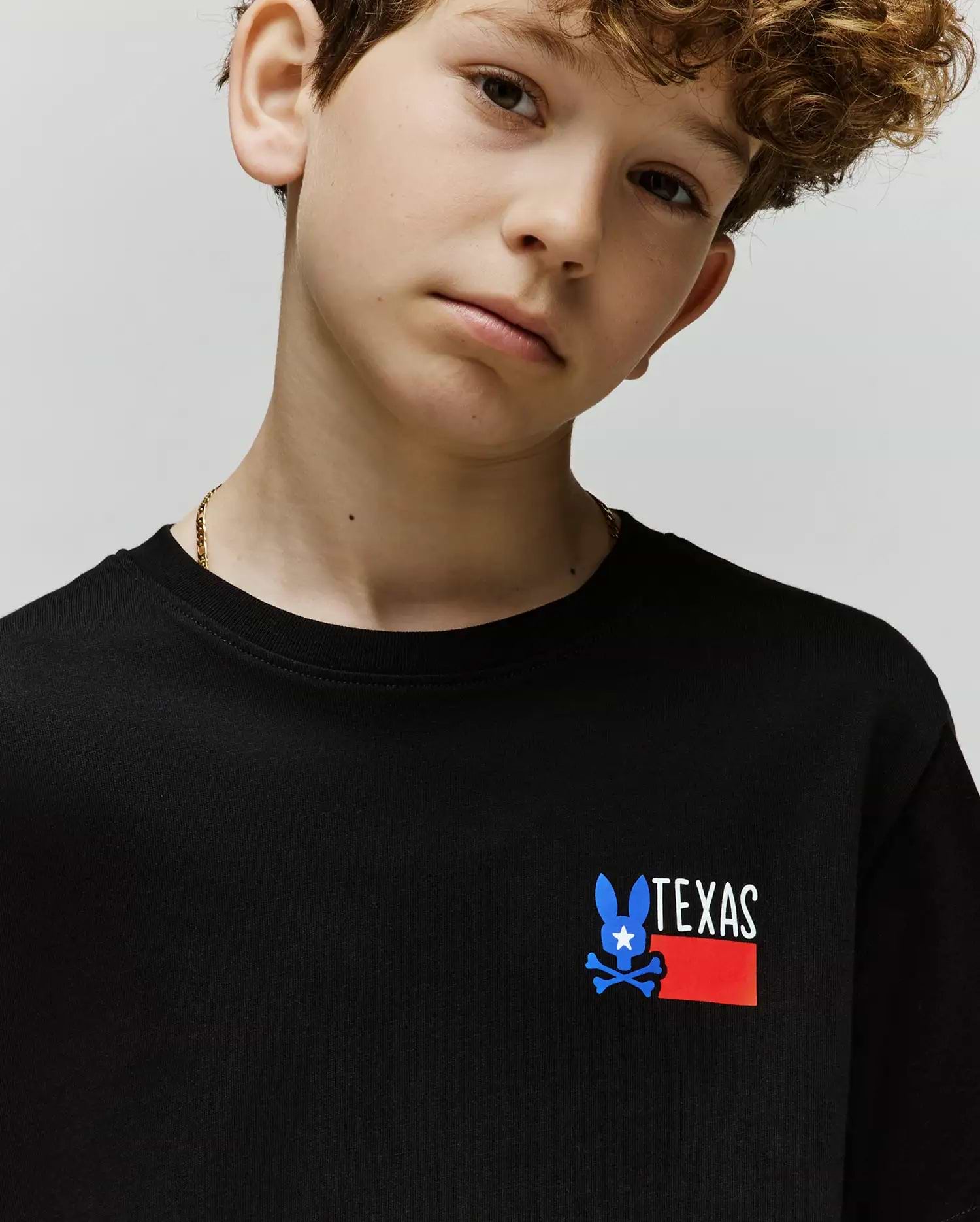 A young boy wears the Psycho Bunny KIDS TEXAS TEE (B0U553W1PC), a black Pima cotton shirt with a blue bunny, white star, and red "TEXAS" graphic on the front, posing calmly against a plain light background.