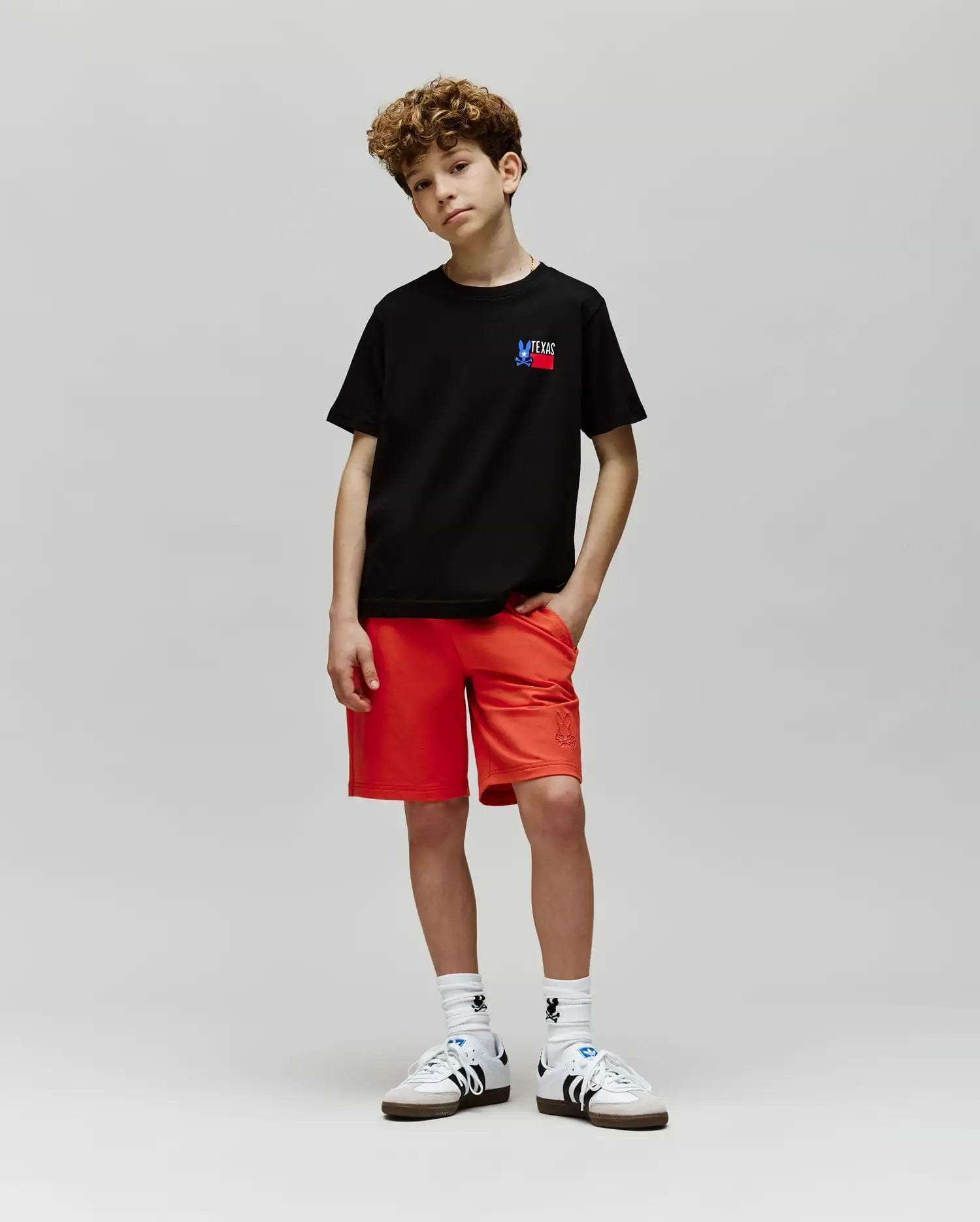 A young boy with curly hair stands against a gray background, wearing Psycho Bunny's KIDS TEXAS TEE (B0U553W1PC) in black, bright red shorts, white socks, and white sneakers with black stripes. One hand is in his pocket and he looks relaxed.