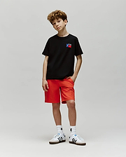 A young boy with curly hair stands against a gray background, wearing Psycho Bunny's KIDS TEXAS TEE (B0U553W1PC) in black, bright red shorts, white socks, and white sneakers with black stripes. One hand is in his pocket and he looks relaxed.