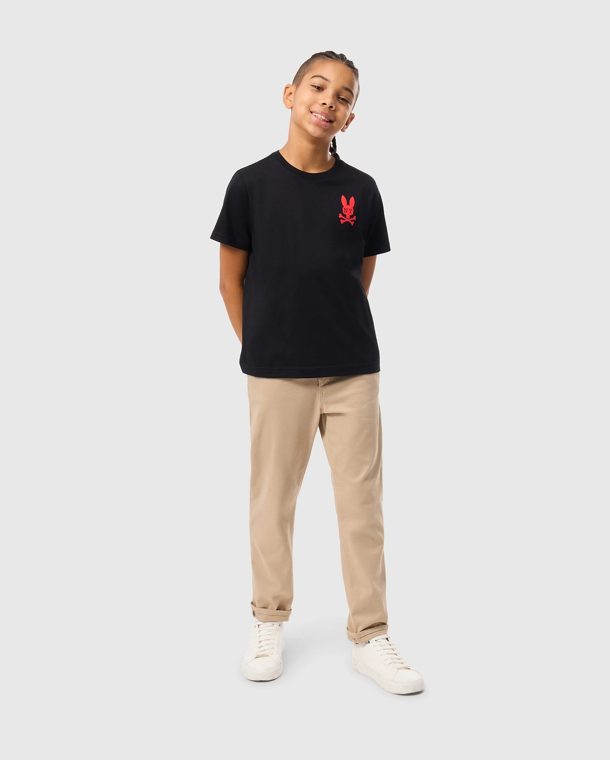 A young boy is smiling with his hands behind his back, wearing a black KIDS NEW YORK CITY TEE by Psycho Bunny that features a red logo on the left chest. He pairs it with beige pants and white sneakers against a plain gray background.