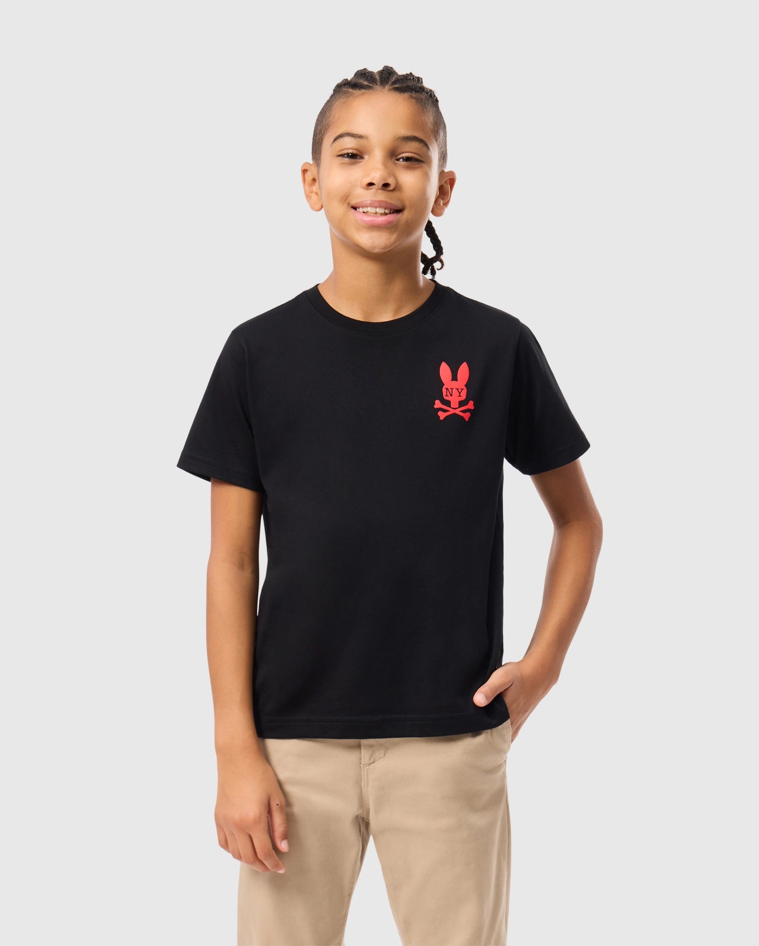 A young person with braided hair smiles while wearing a stylish Psycho Bunny KIDS NEW YORK CITY TEE in black, crafted from soft Pima cotton and featuring a red emblem of a bunny and crossbones. They pair it with beige pants, one hand casually in their pocket, standing against a plain white background.