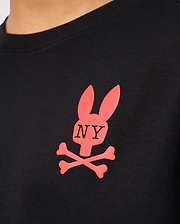 Close-up of a black Psycho Bunny KIDS NEW YORK CITY TEE - B0U554W1PC, crafted from premium Pima cotton and featuring a red logo with a stylized rabbit head above crossed bones. The letters "NY" are printed on the rabbit's face, making this tee both stylish and comfortable for kids.