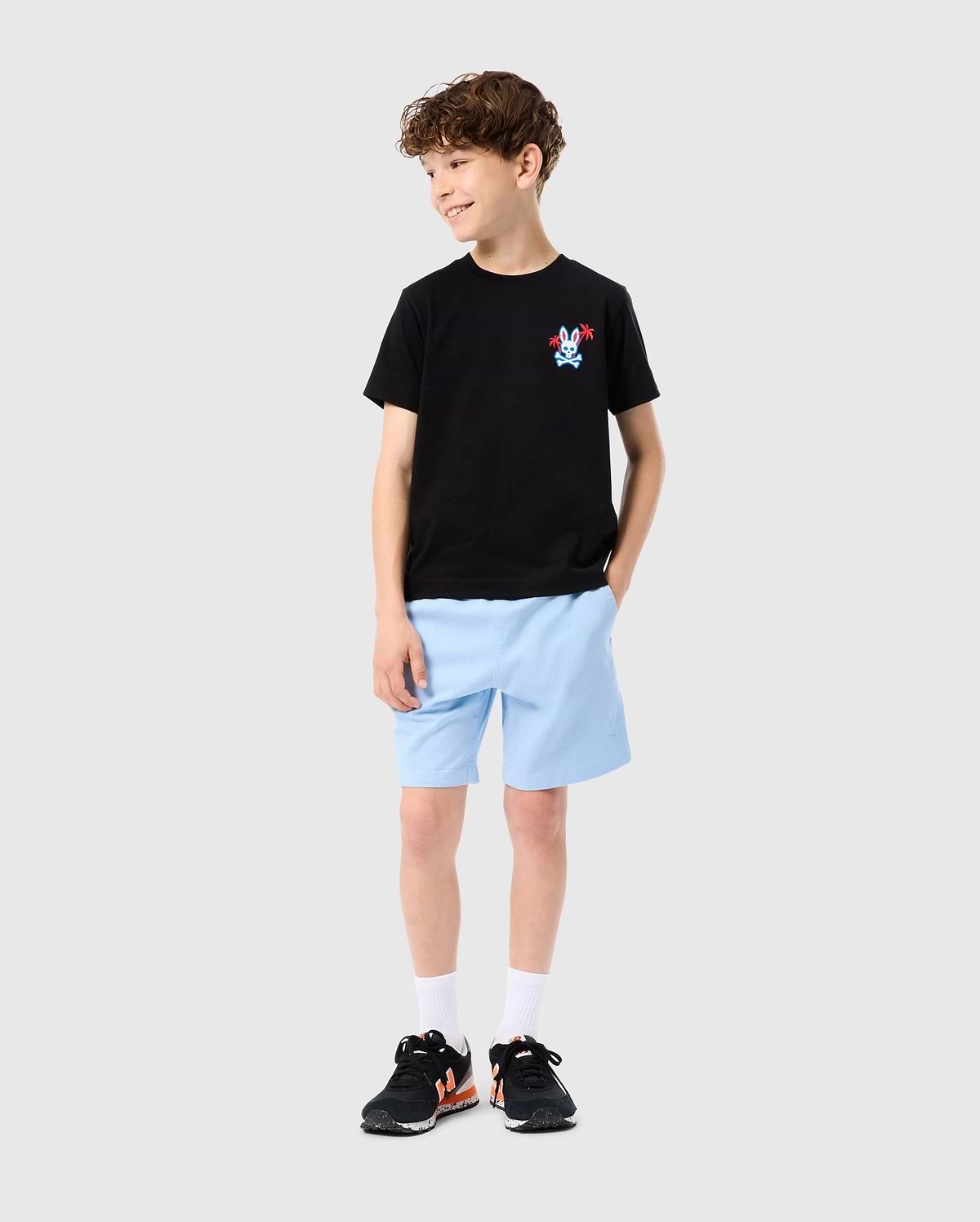 A young boy with curly hair is wearing a Psycho Bunny KIDS LOS ANGELES CITY TEE - B0U861A2PC in black, crafted from Peruvian Pima cotton, adorned with a small red and blue graphic design on the left side. He pairs it with light blue shorts, white socks, and black sneakers. Standing against a plain light grey background, he smiles while looking to his left with one hand in his pocket.