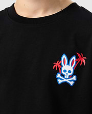 Close-up of the Psycho Bunny KIDS LOS ANGELES CITY TEE - B0U861A2PC in black, made from soft Peruvian Pima cotton. It features a small, colorful graphic on the left side of the chest depicting a bunny skull with crossbones below it, flanked by two small palm trees with red fronds.