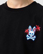Close-up of the Psycho Bunny KIDS LOS ANGELES CITY TEE - B0U861A2PC in black, made from soft Peruvian Pima cotton. It features a small, colorful graphic on the left side of the chest depicting a bunny skull with crossbones below it, flanked by two small palm trees with red fronds.