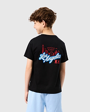A child with curly hair, seen from behind, wears the KIDS LOS ANGELES CITY TEE - B0U861A2PC by Psycho Bunny. The soft black Peruvian Pima cotton tee showcases a vibrant graphic design of buildings and the text "Los Angeles" alongside "Psycho Bunny." Light blue shorts complete the look against a gray background.